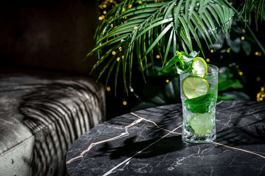 Caipirinha drink alcoholic cocktail served cold in highball glass. Dark luxury lounge bar setting with exotic palm tree shadows on marble table.