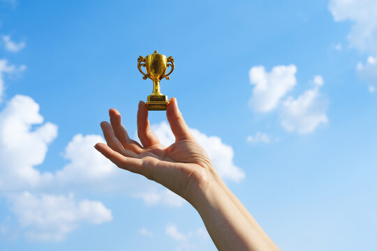 Hand holds and raised small cup trophy, holding cup against clean sky backgrund. Consolation prize award and victory concept.