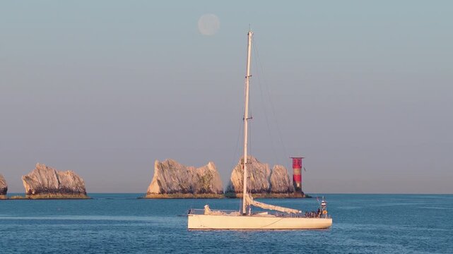 4K panning drone footage of The Needles at sunrise with early morning light, the moon visible in the distance and a sailing yacht in the foreground.