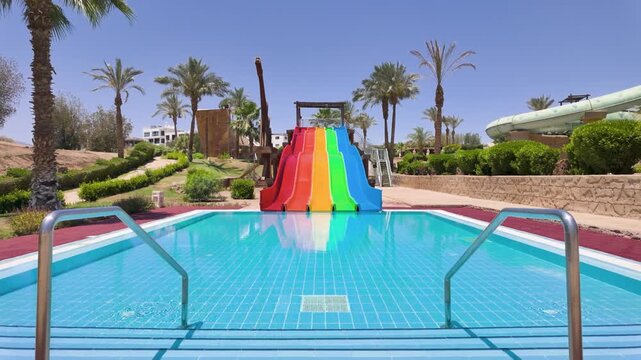 Slow motion footage of a colourful rainbow water slides beside a swimming pool in Sharm El Sheikh Egypt surrounded by palm trees and tropical resort gardens beneath a bright blue sky