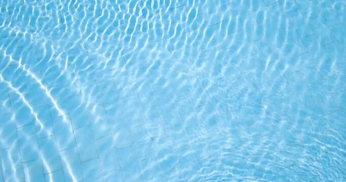Slow motion footage of crystal clear blue swimming pool water gentle ripples reflecting bright sunlight across tiled surfaces creating a relaxing summer holiday background with shimmering patterns