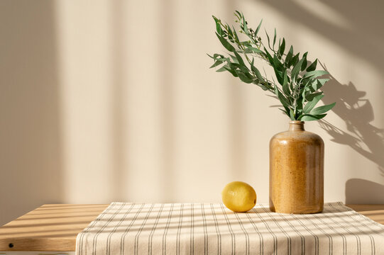 Summer kitchen home interior decor, ceramic vase, lemon on wooden table with stripped linen tablecloth, empty beige wall background with natural sunlight shadows, cottage retro aesthetic
