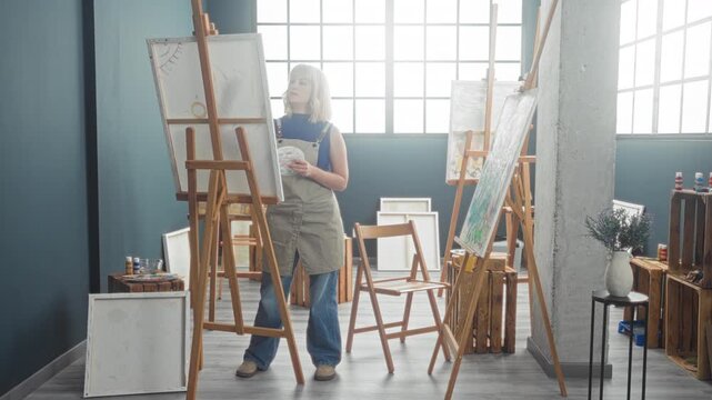 Woman paints on easel holding palette and rag in a studio wearing apron and jeans; quiet concentration creativity craft.