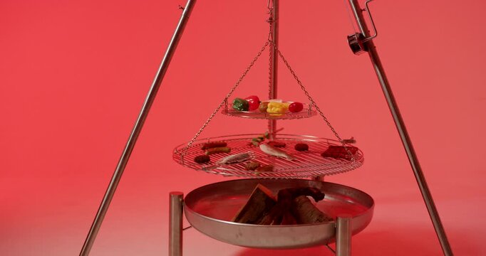 A dual layer circular metal barbecue grill rack rotates over a metal basin containing small logs