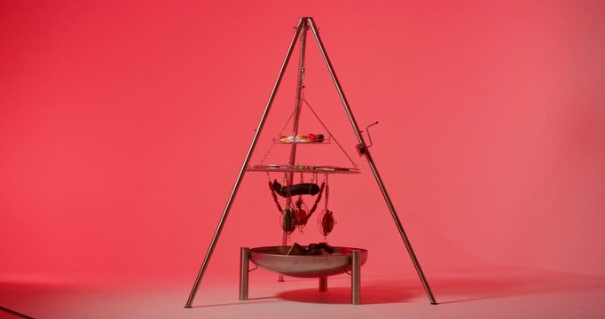 A hanging two tiered barbecue grill rack suspended from a tripod over an empty metal fire pit