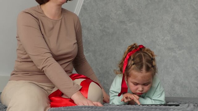 Young woman helping her small daughter look beautiful by arranging her hair and tying a ribbon. Parenting and childhood care concept. Family connection and everyday happiness at home. Mid shot