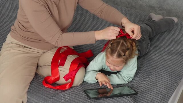 Mother and little daughter enjoying time together while creating a hairstyle with a decorative ribbon. Family leisure and child grooming concept. Positive parenting and warm emotional bonds. Top view