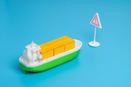 Miniature cargo ship and a triangular warning sign. Maritime hazards, supply chain risks, shipping alerts, trade sanctions concept.