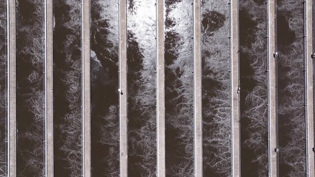 Top down drone shot ascends above active aeration channels at a wastewater treatment facility revealing churning water and foam patterns.