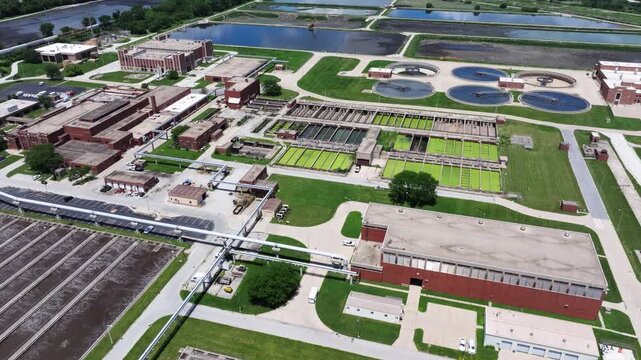 Forward drone shot above a water reclamation facility revealing bright green trickling filter beds, brick operations buildings, circular clarifiers and settling ponds.