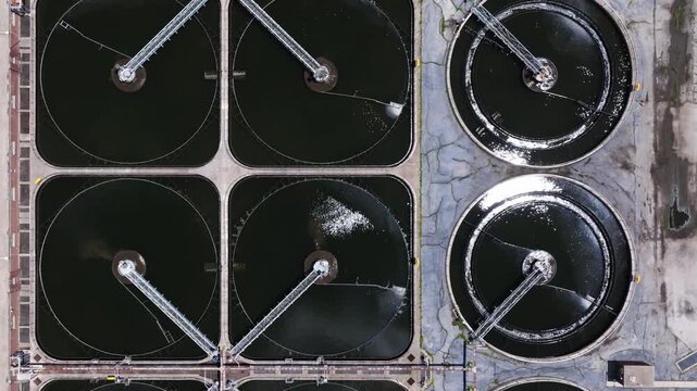 Forward birds eye drone shot above square and circular clarifier tanks with rotating skimmer arms at a wastewater treatment facility.