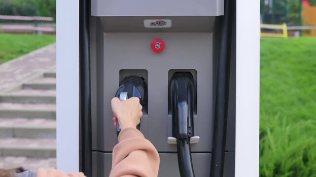 Woman hand places charging plug into station connector during electric vehicle charging process. Charging infrastructure reflects smart mobility, renewable power and environmental education concepts.