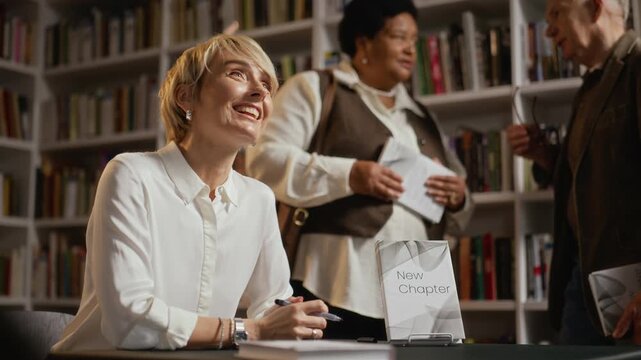 Caucasian adult female author signing books and handing them to readers at book launch event in library. Footage is perfect for promoting literary events or publishing industry marketing