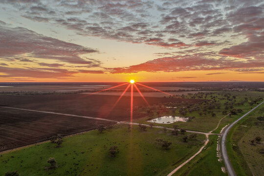 Aerial view of cultivated fields surrounded by a lush green landscape as the rising sun climbs above the horizon, illuminating a colourful morning sky and the peaceful countryside in NSW, Australia.