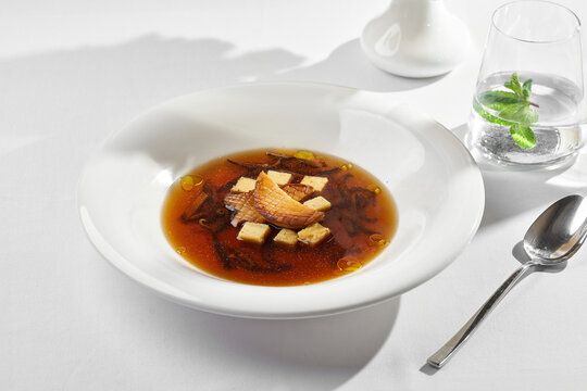 Gourmet mushroom consomme soup with grilled king oyster mushrooms, croutons, and a glass of mint water on a sunny restaurant table.