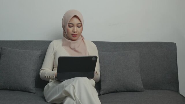 Serious young Asian woman wearing hijab is working using her tablet device while sitting down on a sofa.
