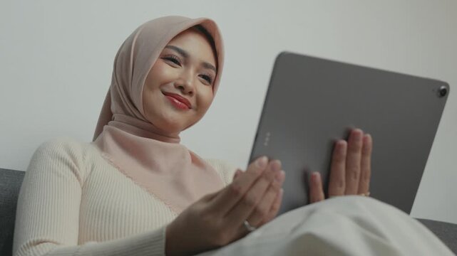 Happy young Asian Muslim woman wearing hijab is using tablet device while sitting on a sofa.