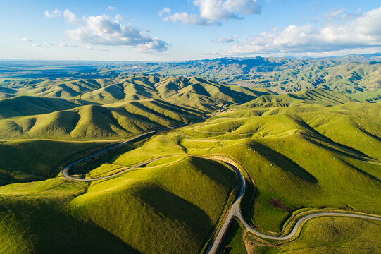 Aerial view of a winding road cutting through vibrant green rolling hills under a bright blue sky in Kern County, California, United States.