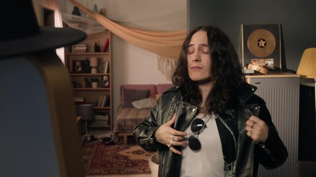 Young rocker with long hair putting on leather jacket, trying on black hat and sunglasses in front of mirror. Clip for fashion retail marketing and youth lifestyle commercials