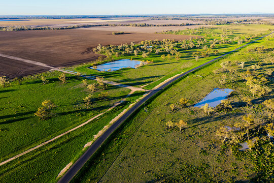 Aerial scene showcasing cultivated farmland with a rural dirt road connecting to a main road, scattered farm dams and a lush green landscape near Pilliga, highlighting the striking patterns, textures
