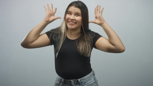 Hispanic young woman shows open hands by face, smiling with tongue out and wearing black top in studio; playful joy.