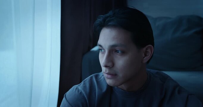 Alone man sit floor by window at night. Sad young person looking out in dark room at home. Gloomy atmosphere, loneliness, depression, solitude. Melancholy mood, introspection, psychological struggle.
