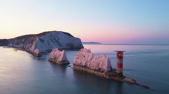 Panning drone shot revealing the Needles lighthouse, white chalk stacks, and cliffs on the Isle of Wight under a golden pre-sunrise dawn sky. Calm morning 4K aerial.