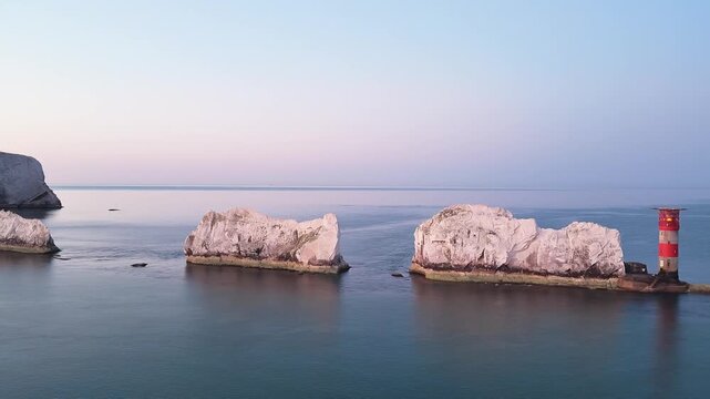 Panning drone shot revealing the Needles lighthouse, white chalk stacks, and cliffs on the Isle of Wight under a golden pre-sunrise dawn sky. Calm morning 4K aerial.