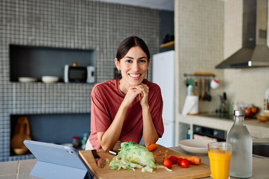 Happy young latina woman having fun preparing food and looking for recipes online using a tabletor laptop in kitchen