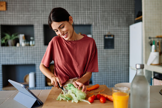 Happy young latina woman having fun preparing food and looking for recipes online using a tabletor laptop in kitchen