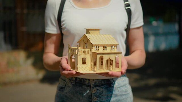Closeup wooden house model in woman hands. New apartment purchase and mortgage financing concept with relocation planning and residential investment lifestyle. Urban background supporting dream home