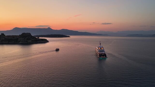High-angle aerial drone shot of the massive luxury mega yacht Al Mirqab anchored in the sea near Corfu Old Fortress during a vibrant golden hour sunset, Greece.