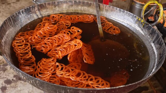 Close-up of golden orange jalebi sweets absorbing sweet syrup in a large commercial cooking pan. Traditional crispy jalebi by soaking the fried spirals in sweet sugar syrup at a village.