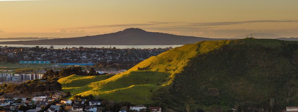 Auckland, New Zealand. The sun sets over the rolling green hills of Mt Wellington and suburban landscape, casting long shadows and a warm glow. Rangitoto in the distance.