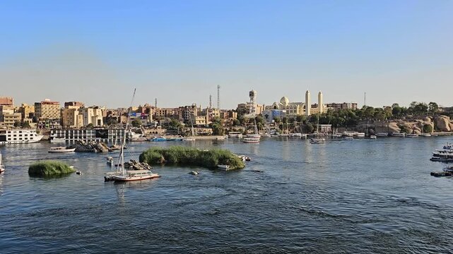 Scenic Nile River view from Elephantine Island at Aswan, Egypt, with tourist ferry boats, granite rocks, lush green island, sandy desert hills and city under blue sky.