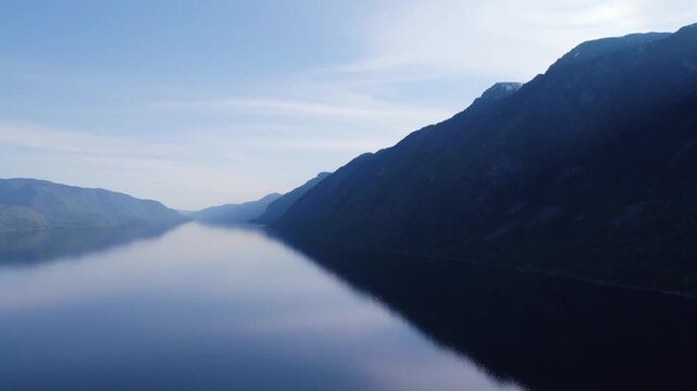 Dense fog rolling across fjord valley beside steep dark mountain silhouette with mirror smooth water, pale blue sky and distant ridgelines in quiet scenic northern landscape