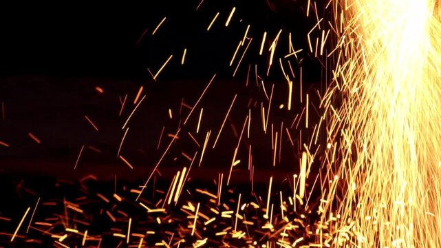 Bright yellow and orange fiery sparks showering down and scattering across a dark surface during an industrial metalworking process.