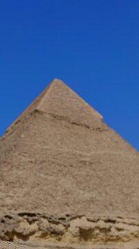 Pyramid of Khafre framed between large granite boulders with desert plateau and camels at Giza, Egypt. Unique perspective of ancient wonder under vivid blue sky.