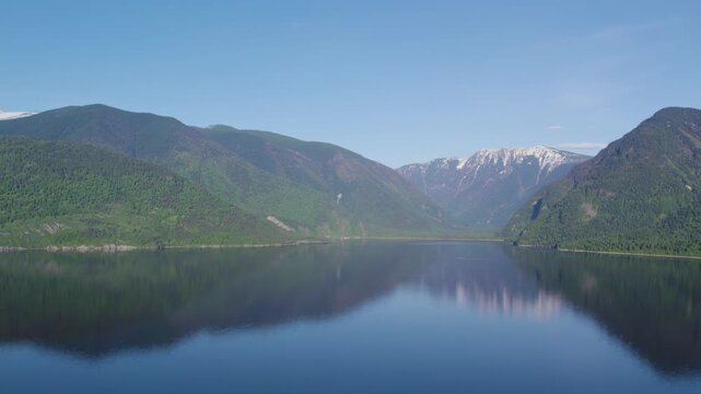 Tranquil fjord panorama with mirror like water, gentle central ripples, forested mountains, distant snow capped summit, clear blue sky and remote alpine wilderness
