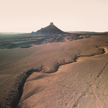 Aerial view of Factory Butte rising above the arid, eroded desert landscape of the badlands with intricate drainage patterns and soft golden light in Wayne County, Utah, United States.