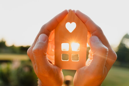 Two hands gently hold a wooden house model with a heart shape while the sun shines through the window. Loving family home and real estate mortgage concept.