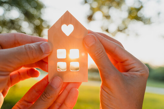 Hands hold a small wooden house silhouette as the warm setting sun brightly shines through its square window. New home ownership and hopeful real estate concept.