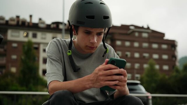 A teenage boy wearing a protective helmet sits in a skatepark and focuses on using his mobile phone.