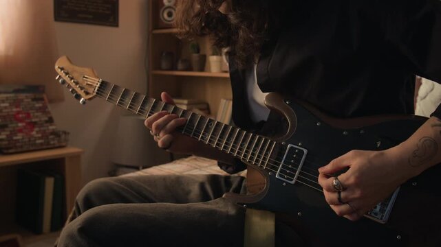 Midsection shot of young man with long hair adjusting knobs and playing electric guitar while sitting on bed. Clip for music education ads or youth lifestyle campaigns