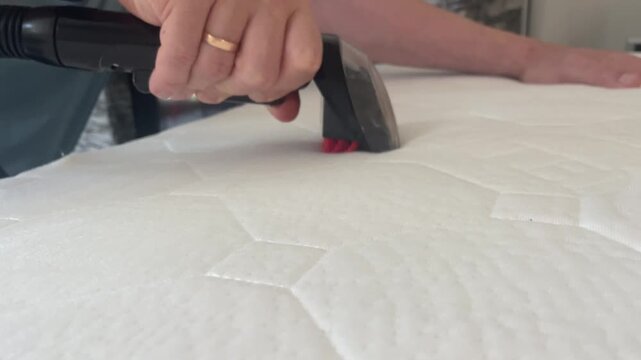Horizontal close up shot of a person deep cleaning and steaming a white fabric mattress surface with a handheld steam cleaner nozzle tool for hygiene home maintenance. High quality 4k footage