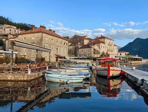 Perast port framed by baroque stone palaces and seafront promenade with moored fishing boats reflecting on calm Boka Kotorska waters at golden hour. Warm light bathes terracotta roofs and quay
