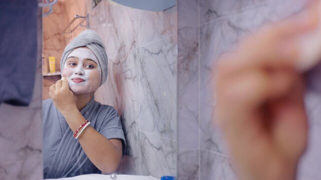 Indian woman removing dried face pack with soft cotton pad in front of a mirror, dressed in bathrobe in the bathroom.