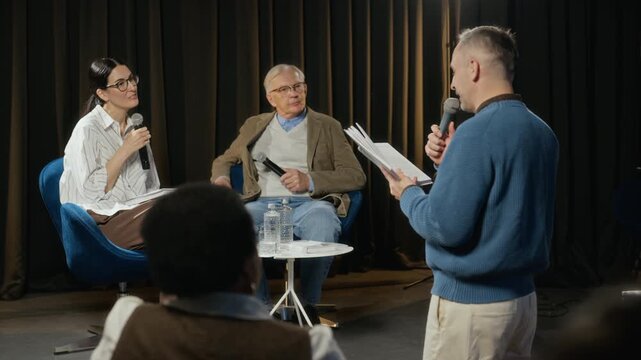 Middle-aged Caucasian man holding microphone and reading book, presenting to panel of young adult Caucasian woman and senior Caucasian man. Clip is ideal for corporate communication videos