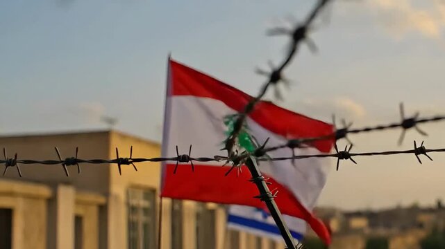 the national flag of lebanon waves in the wind against the sky, the flag of israel is visible below through rows of barbed wire, symbolizing border conflict