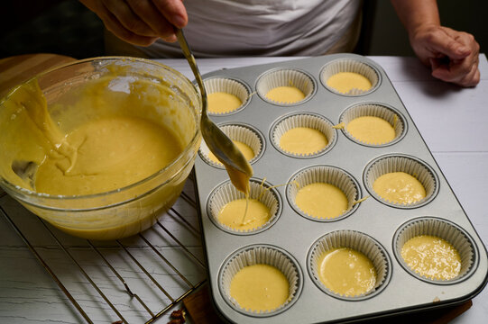 Hand pours golden custard into paper cups. White apron suggests home baking or dessert making. Spoon drizzles smooth, warm liquid with care and precision. Cupcake liners wait on cooling rack beneath